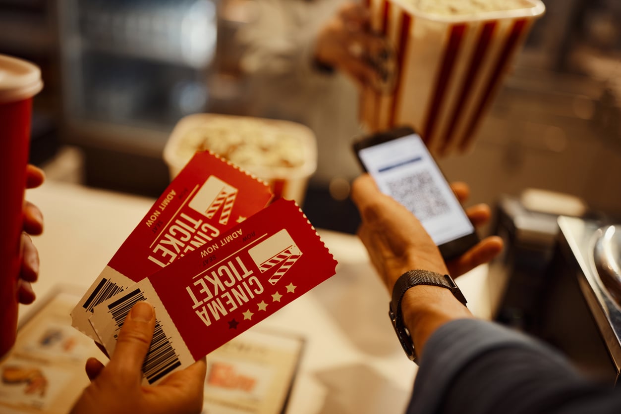 Two hands at a concession stand: one holding two red cinema tickets and a large popcorn, the other showing a QR code on a smartphone for scanning—if only these treats were at 1991 prices. A large drink cup sits on the counter.