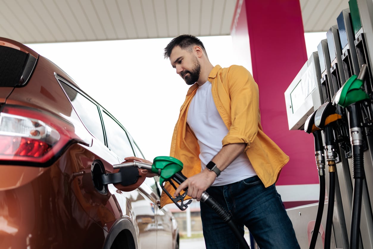 A man in a yellow shirt and white t-shirt is standing at a gas station, holding a green fuel nozzle while refueling his brown SUV, perhaps wishing for 1991 prices. The gas pumps and canopy roof are visible in the background.