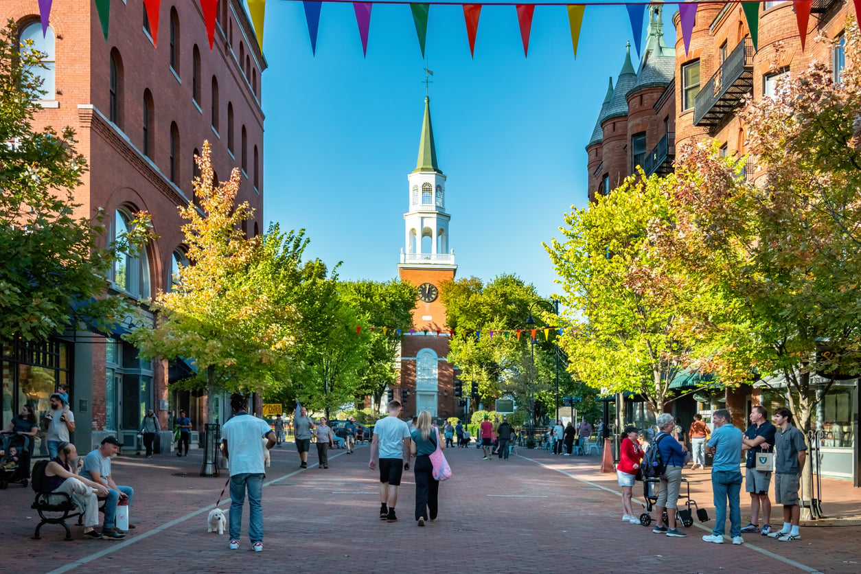 People stroll along a lively brick-paved pedestrian street in one of America’s most charming affordable college towns, lined with trees and historic red brick buildings, colorful pennant banners overhead, and a white-steepled church under a clear blue sky.