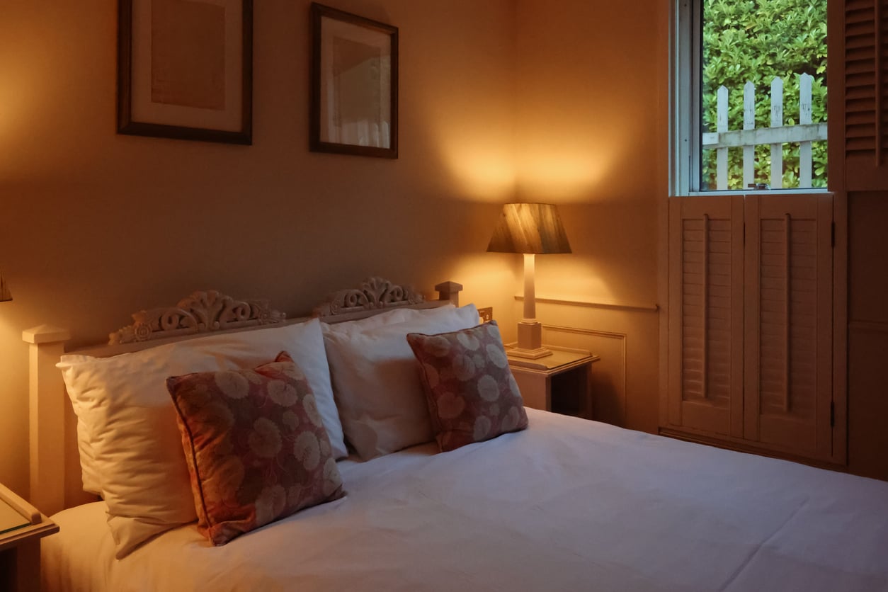A cozy bedroom with a double bed, two patterned pillows, warm lamp lighting, framed art on the wall, and a window with shutters partly open to a view of greenery and a wooden fence outside.