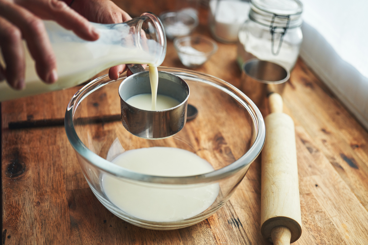A person pours milk from a glass bottle into a metal measuring cup over a clear glass bowl on a wooden table. Nearby are a rolling pin, jars, and other baking tools.