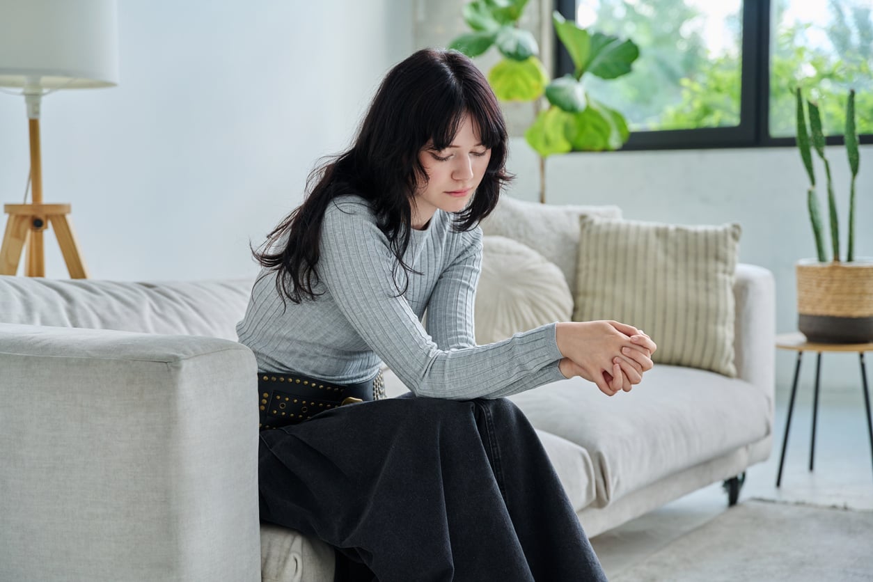 A woman with long dark hair sits on a light-colored sofa in a bright living room, looking down thoughtfully with her hands clasped—perhaps reflecting on delayed purchases. Green plants and large pillows are visible in the background.