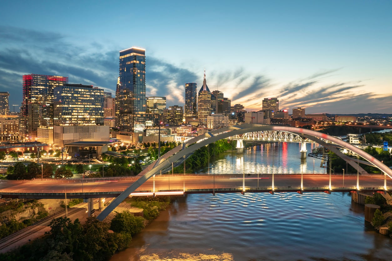 City skyline at sunset with tall buildings, a lit arched bridge spanning a river, and reflections on the water—offering one of the stunning Florida alternatives for an evening view. The sky is partly cloudy with fading daylight.