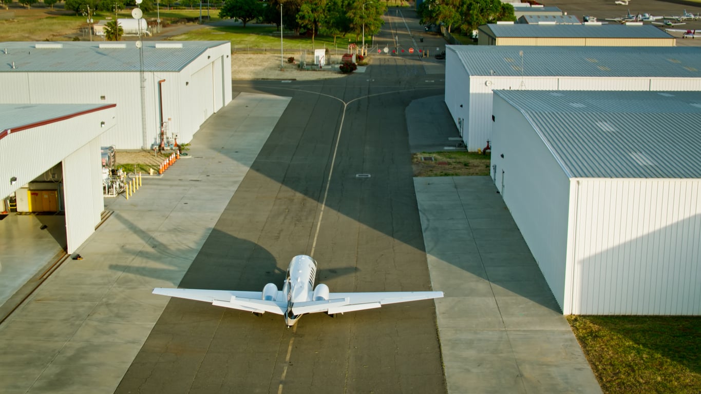 A small white airplane is taxiing on a paved runway between large white hangars at an airport, with green grass and trees visible in the background.