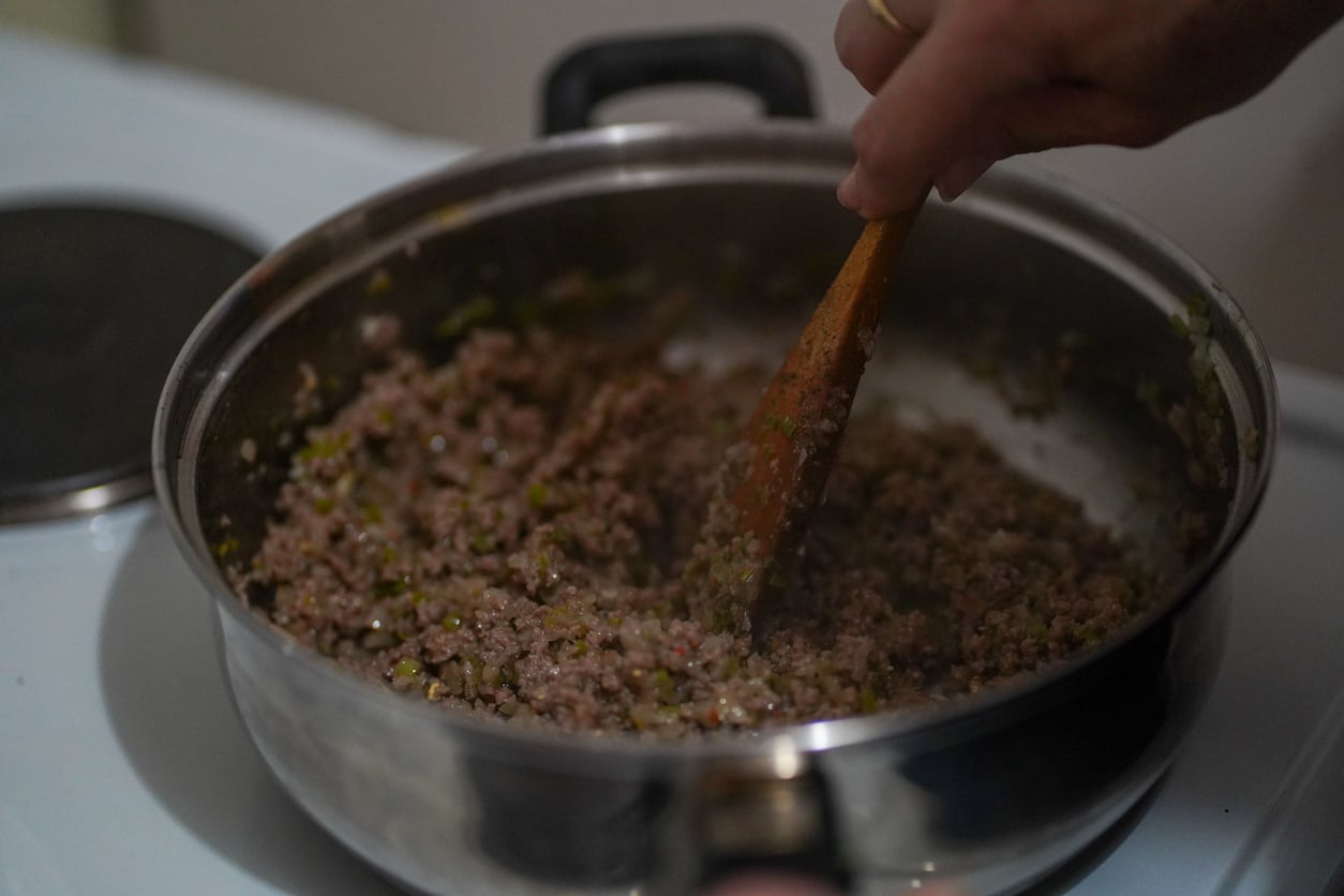 A hand stirs ground meat and chopped vegetables in a stainless steel pan with a wooden spoon on a stovetop, creating a tasty homemade meal without relying on overpriced products.