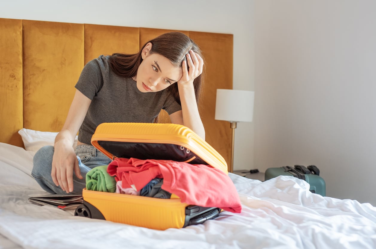 A young woman sits on a bed in a hotel room, looking frustrated as she tries to close an overstuffed yellow suitcase filled with colorful clothes. A lamp and another suitcase are visible, highlighting the importance of hotel room safety while traveling.