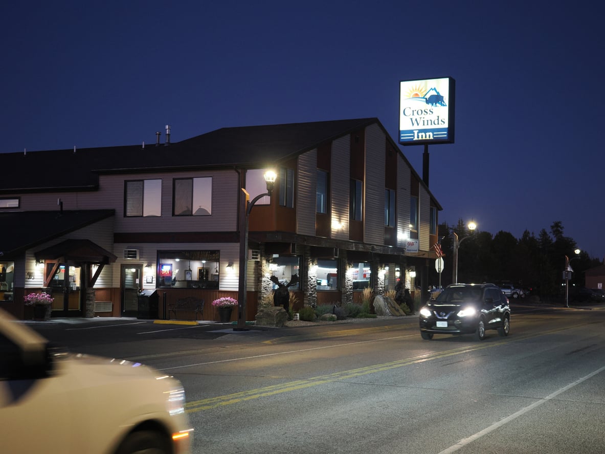 A street view at dusk shows cars passing by the Cross Winds Inn, a two-story building with lit windows and a bright sign advertising classic 1991 prices, under a clear evening sky.