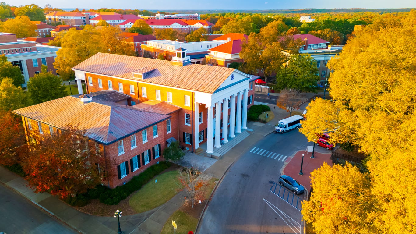 Aerial view of a red-brick university building with white columns, nestled in one of the most affordable college towns, surrounded by autumn foliage and parked cars, under a clear sky during golden hour.