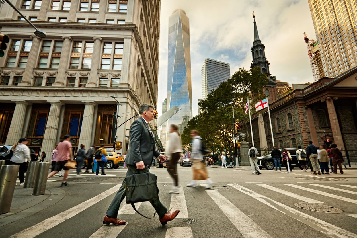 A man in a suit carries a bag while crossing a busy city street at a crosswalk, surrounded by blurred pedestrians and tall buildings, embodying the dynamic spirit of Small Business States with a skyscraper rising in the background.