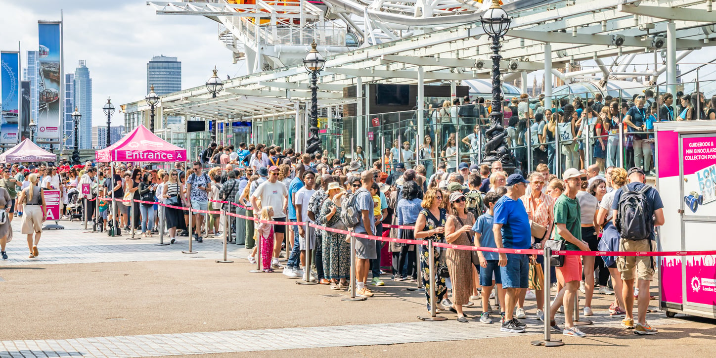 A large crowd of people stands in long, organized lines outside a glass building on a sunny day, with pink “Entrance A” signs and red barriers guiding the queues—perhaps hoping theme park hacks will help them get inside faster. Tall city buildings rise in the background.