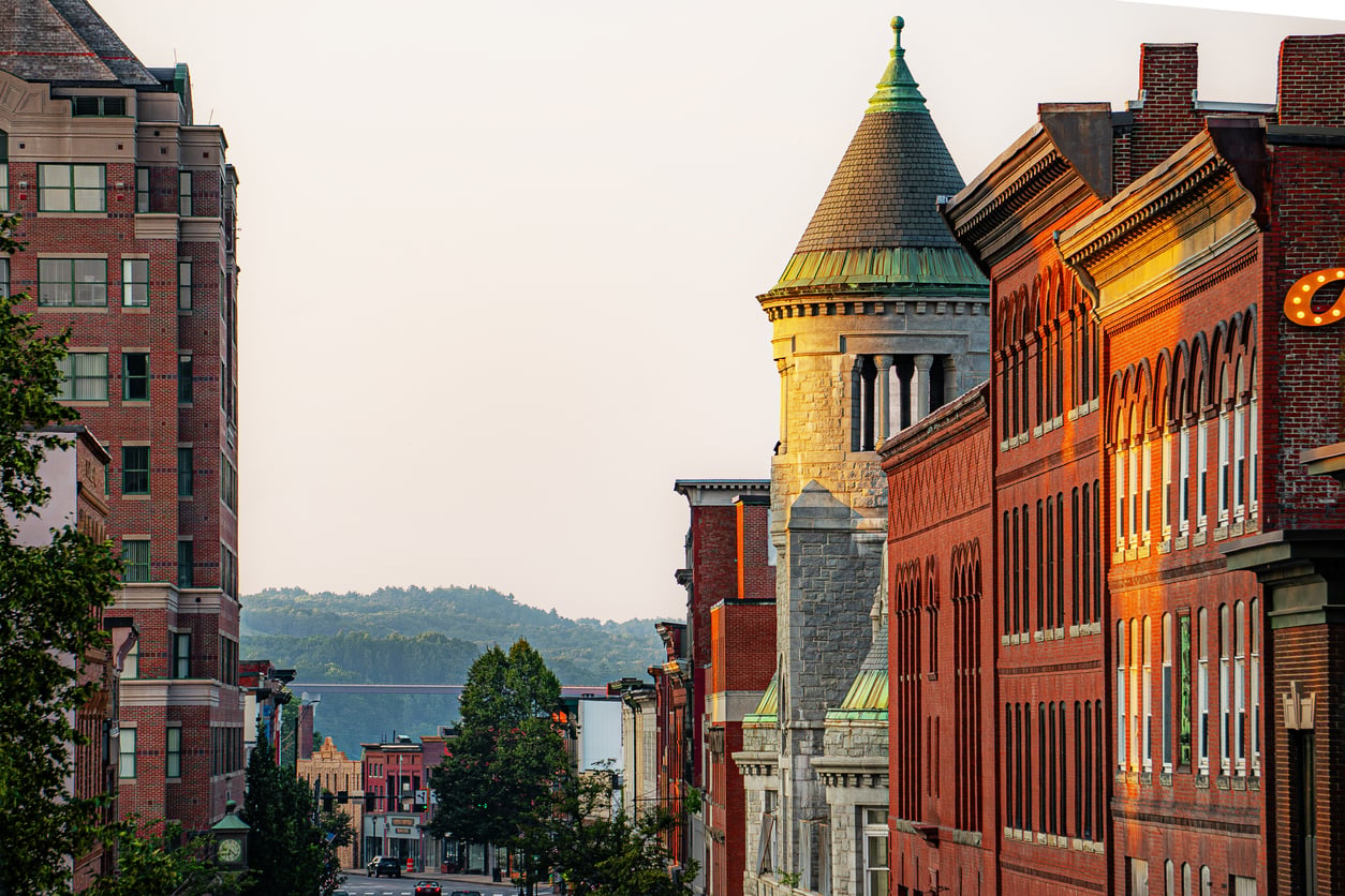 Downtown street lined with historic red brick buildings and a stone tower, bathed in warm sunlight, with green hills visible in the background under a clear sky.
