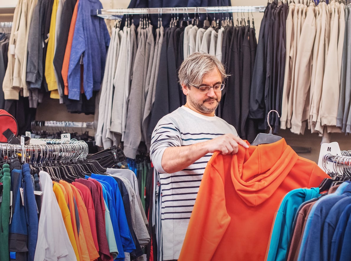 A man with glasses and gray hair examines an orange hoodie in a clothing store, surrounded by racks of various colored shirts, pants, and hoodies—perhaps considering products men skip buying.