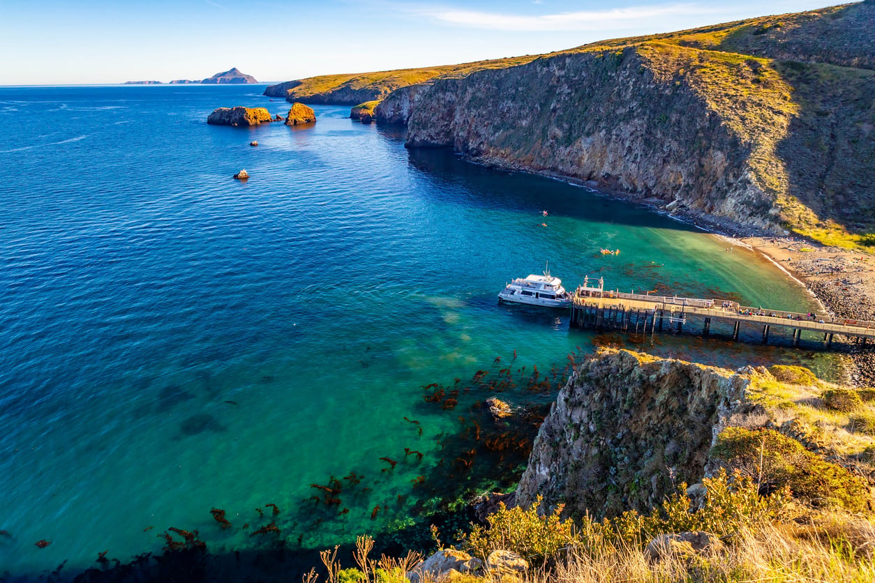 A docked boat at a wooden pier extends into a small, clear blue bay surrounded by rocky cliffs and green hills, with distant islands visible under a sunny sky.