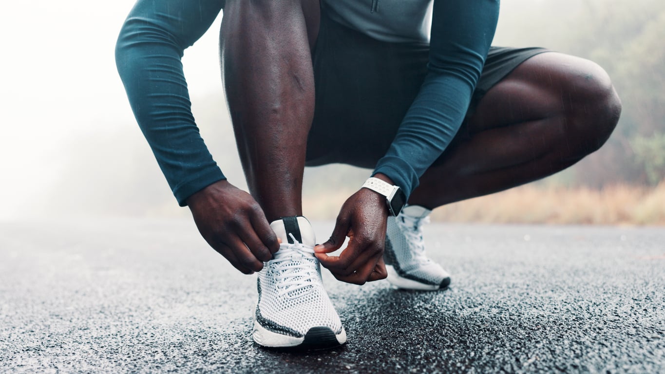 A person in athletic wear crouches on a wet road, tying the laces of their white running shoes—classic products men still buy. The misty background reveals blurred trees and grass, adding to the fresh, active atmosphere.