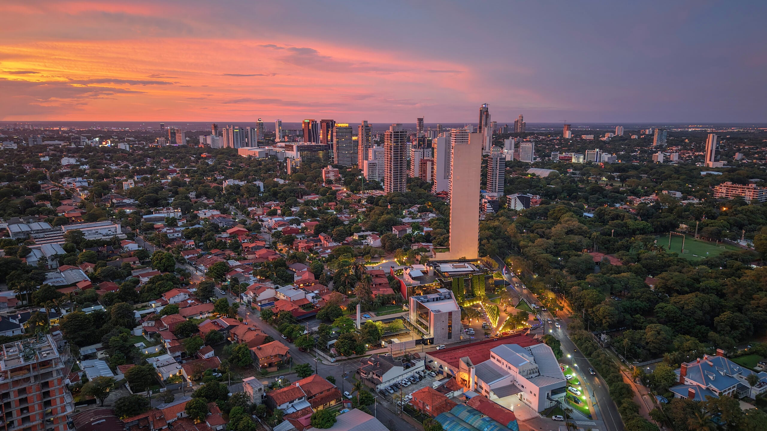 Aerial view of a city at sunset, featuring a mix of tall modern buildings and residential houses surrounded by trees, with a colorful sky in the background.