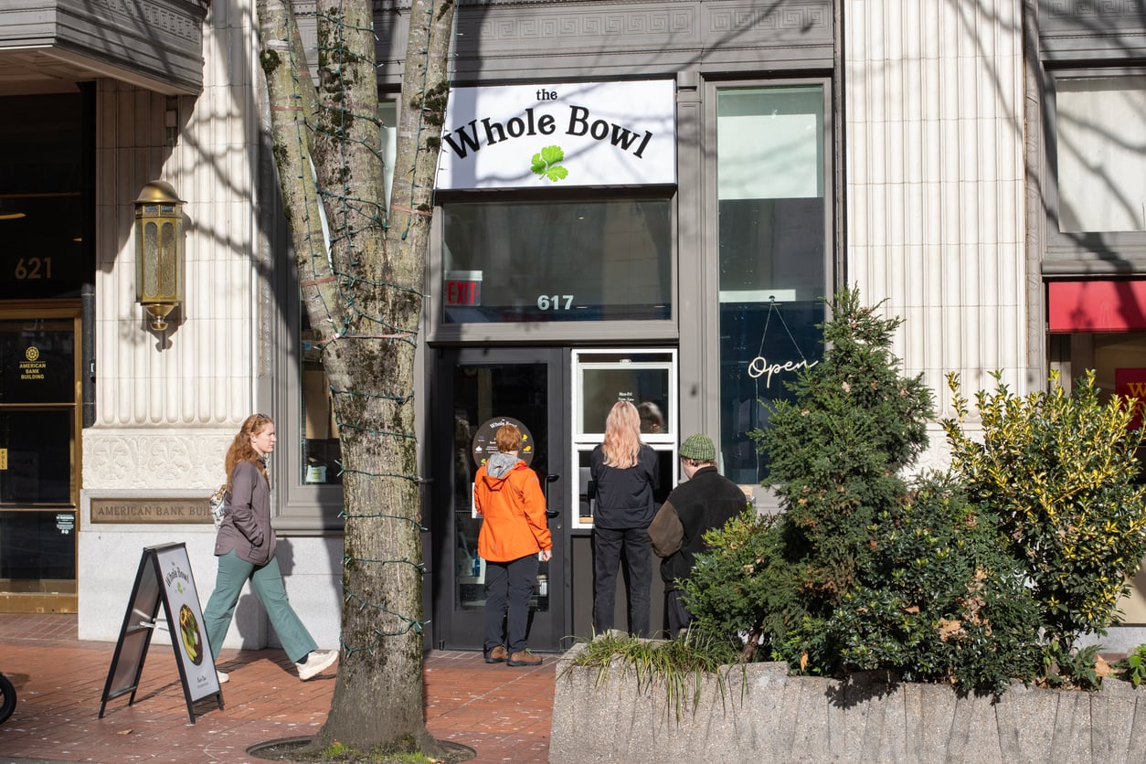 Three people stand outside The Whole Bowl restaurant, a vibrant spot highlighted in Small Business States, on a sunny sidewalk; one enters as two wait by the door. A tree and planter with shrubs enhance the welcoming scene.