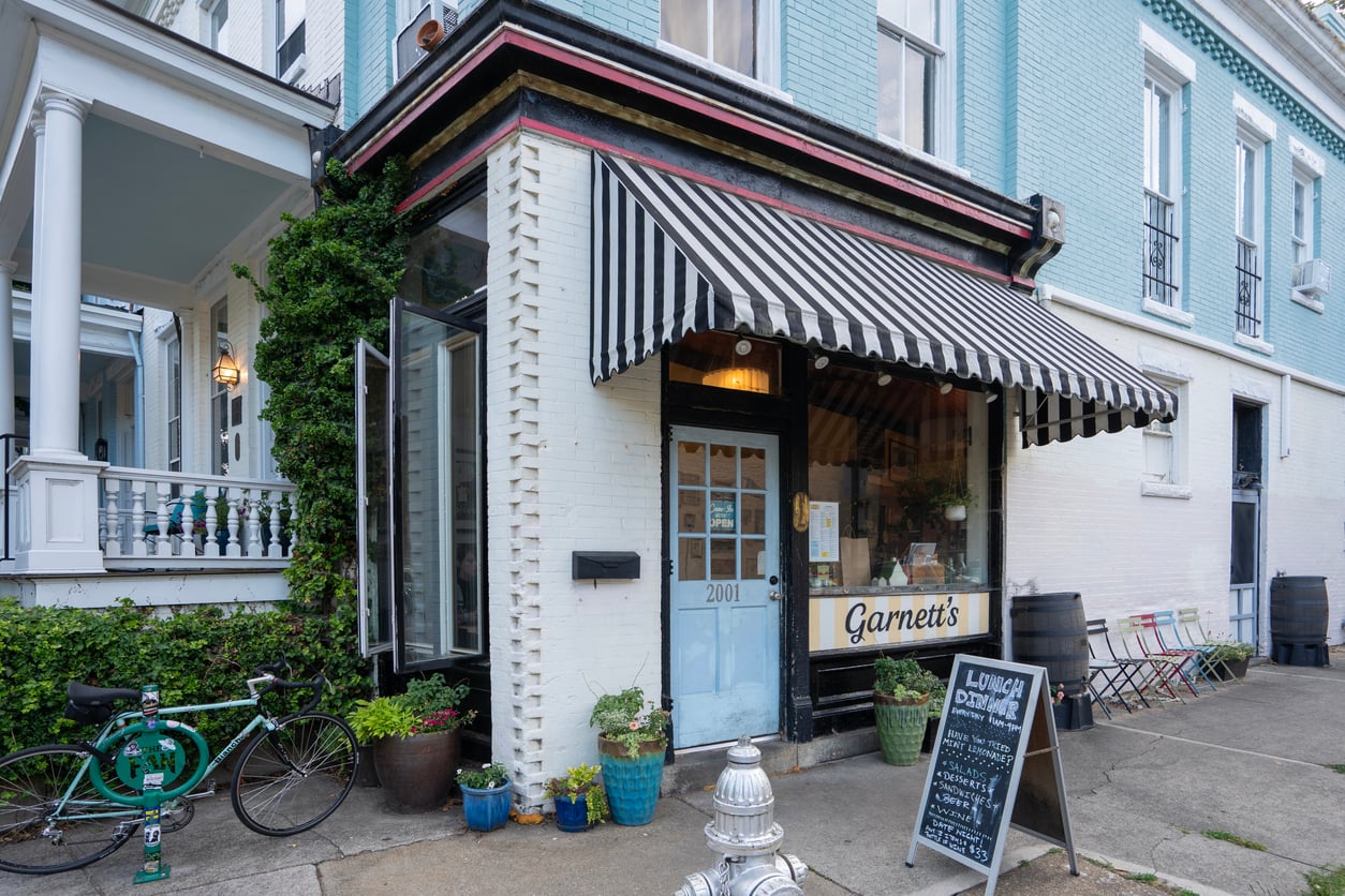 A corner café with a black-and-white striped awning welcomes guests—an inviting small business staple for locals and visitors alike. Potted plants, ivy-covered walls, and a green bicycle capture the charm found across Small Business States.