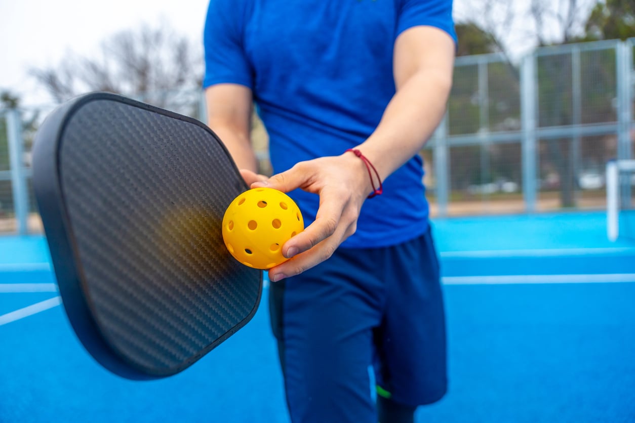 A person in blue sportswear holds a yellow perforated pickleball and a black paddle on a blue outdoor court, enjoying one of the most popular budget-friendly hobbies.