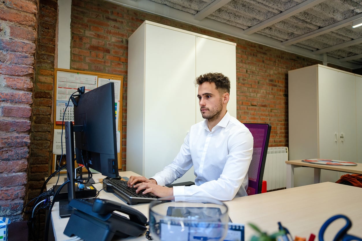A man in a white shirt sits at a desk in a modern office, working on a desktop computer. The workspace features brick walls, cabinets, and office supplies.