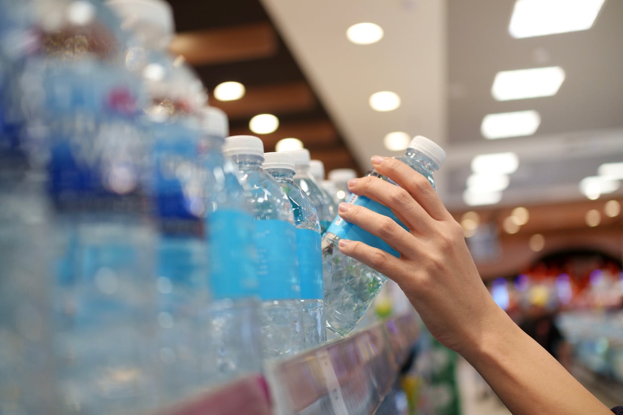 A person’s hand selecting a plastic water bottle from a shelf stocked with multiple overpriced products in a brightly lit store.