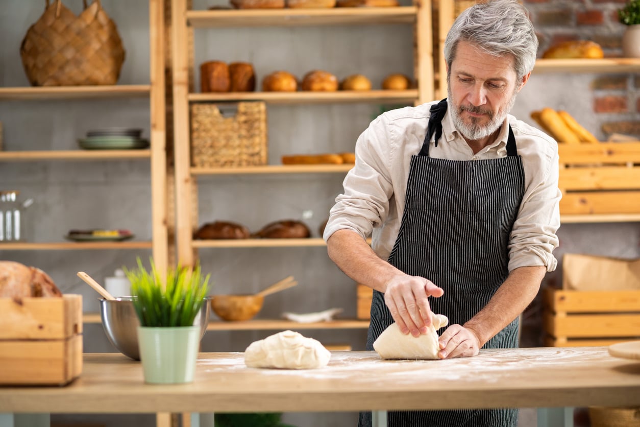 A man wearing an apron kneads dough on a floured wooden table in a bakery with shelves of bread and pastries in the background, showcasing baking as one of many budget-friendly hobbies.