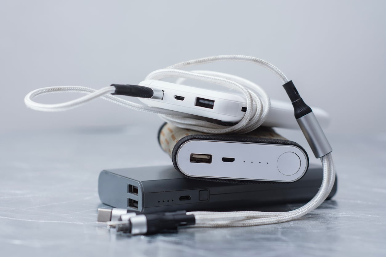 Three portable power banks are stacked on a gray surface, with two USB charging cables draped over them. Perfect for theme park hacks, these power banks feature visible USB ports and LED indicator lights to keep your devices charged all day.