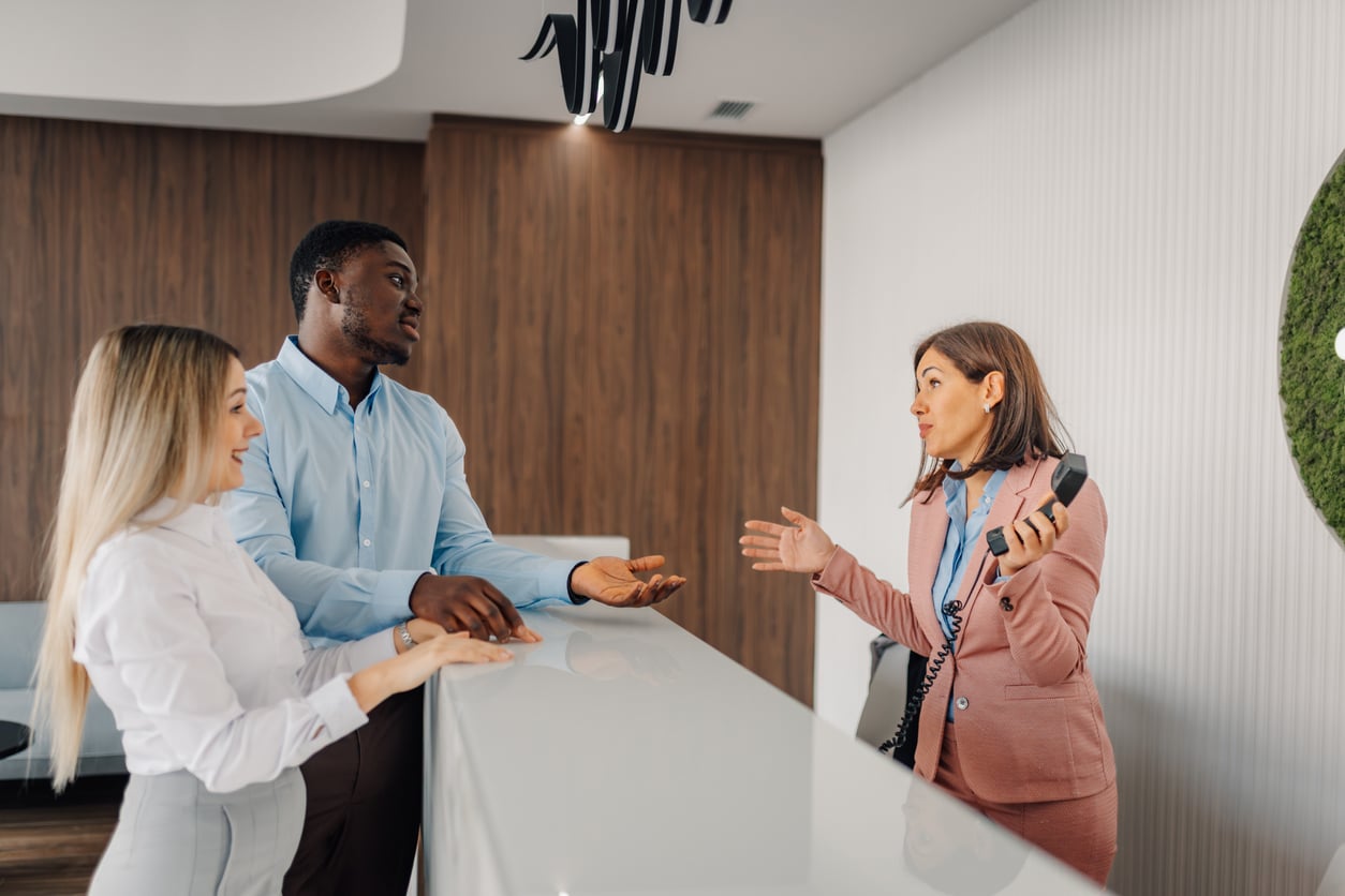 Three people stand at a reception desk; two guests, a man and a woman, talk with a female receptionist in a pink blazer, who gestures expressively while holding a key card—showcasing the welcoming service of future Hotel Brands 2026.