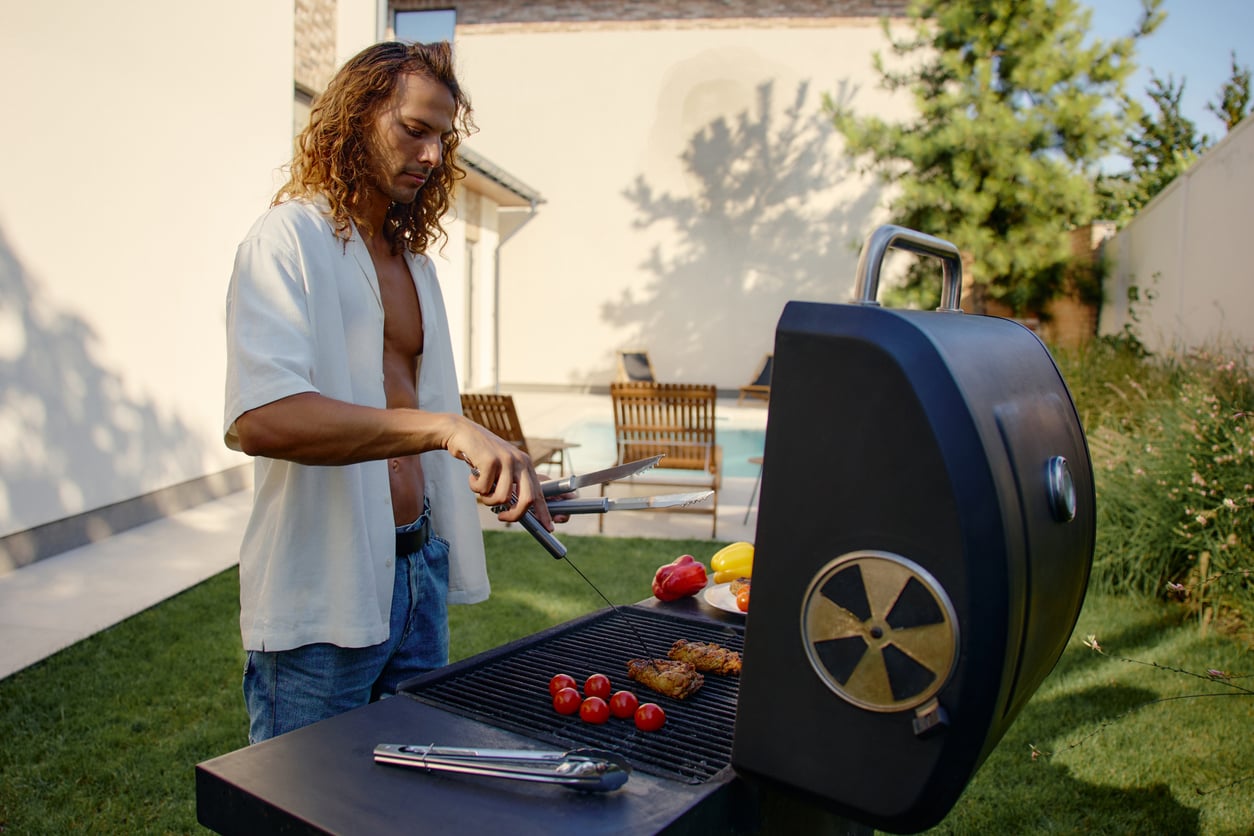 A man with long hair grills assorted vegetables and meat on a barbecue in a backyard, using tongs—classic tools among products men still buy. There’s a pool and lounge chairs in the background on a sunny day.