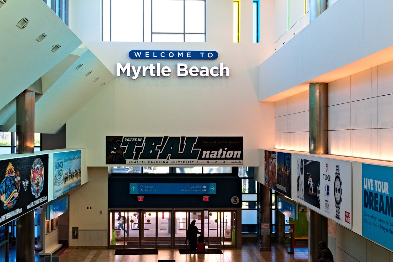 A bright airport terminal features a large "Welcome to Myrtle Beach" sign above the entrance, with various advertisements and banners displayed along the walls. Natural light streams in from high windows.