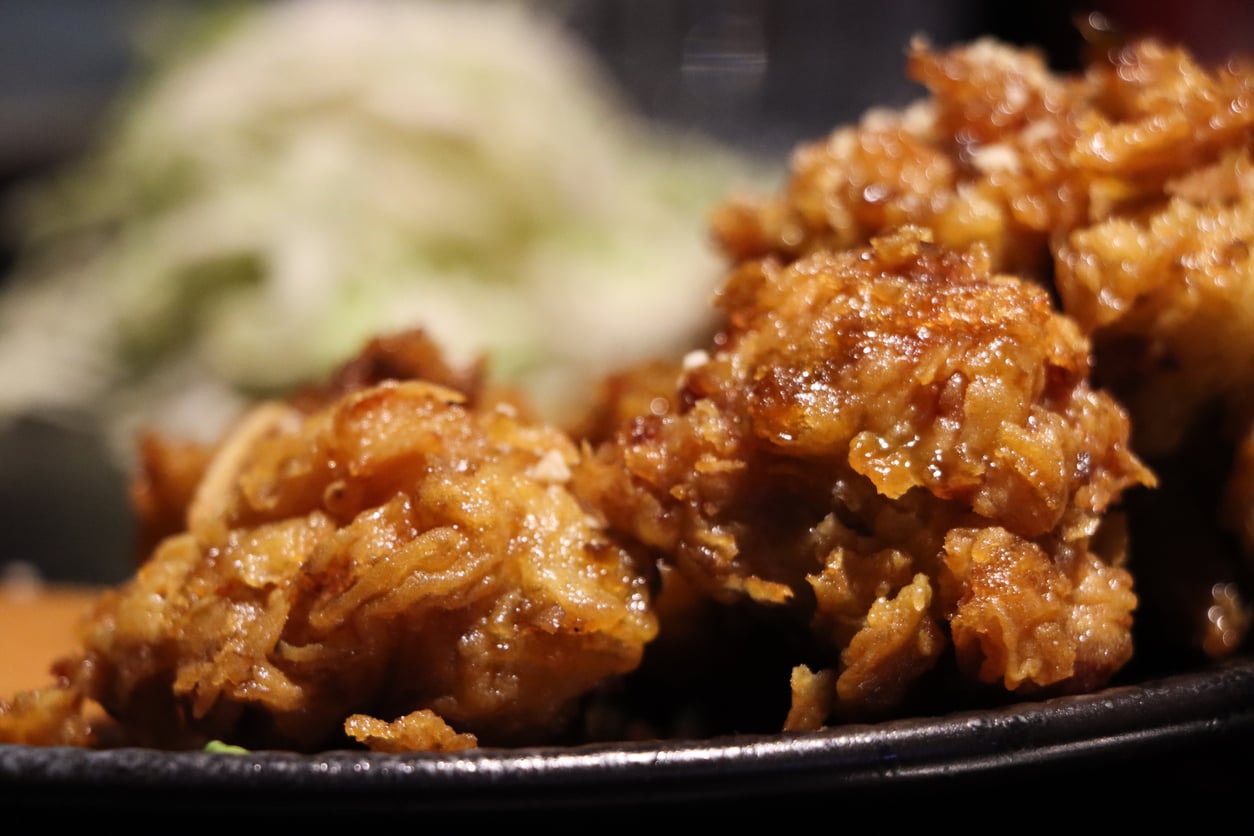 Close-up of crispy, golden-brown fried chicken pieces on a black plate, with a blurry background of shredded cabbage or lettuce.