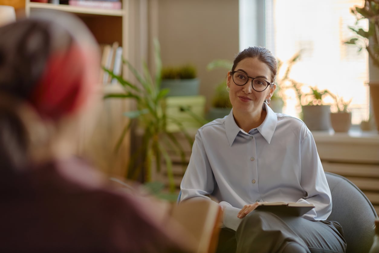 A woman in glasses and a light blue shirt sits with a notepad, attentively listening to another person wearing a headscarf in a cozy, plant-filled office during a mental health session.