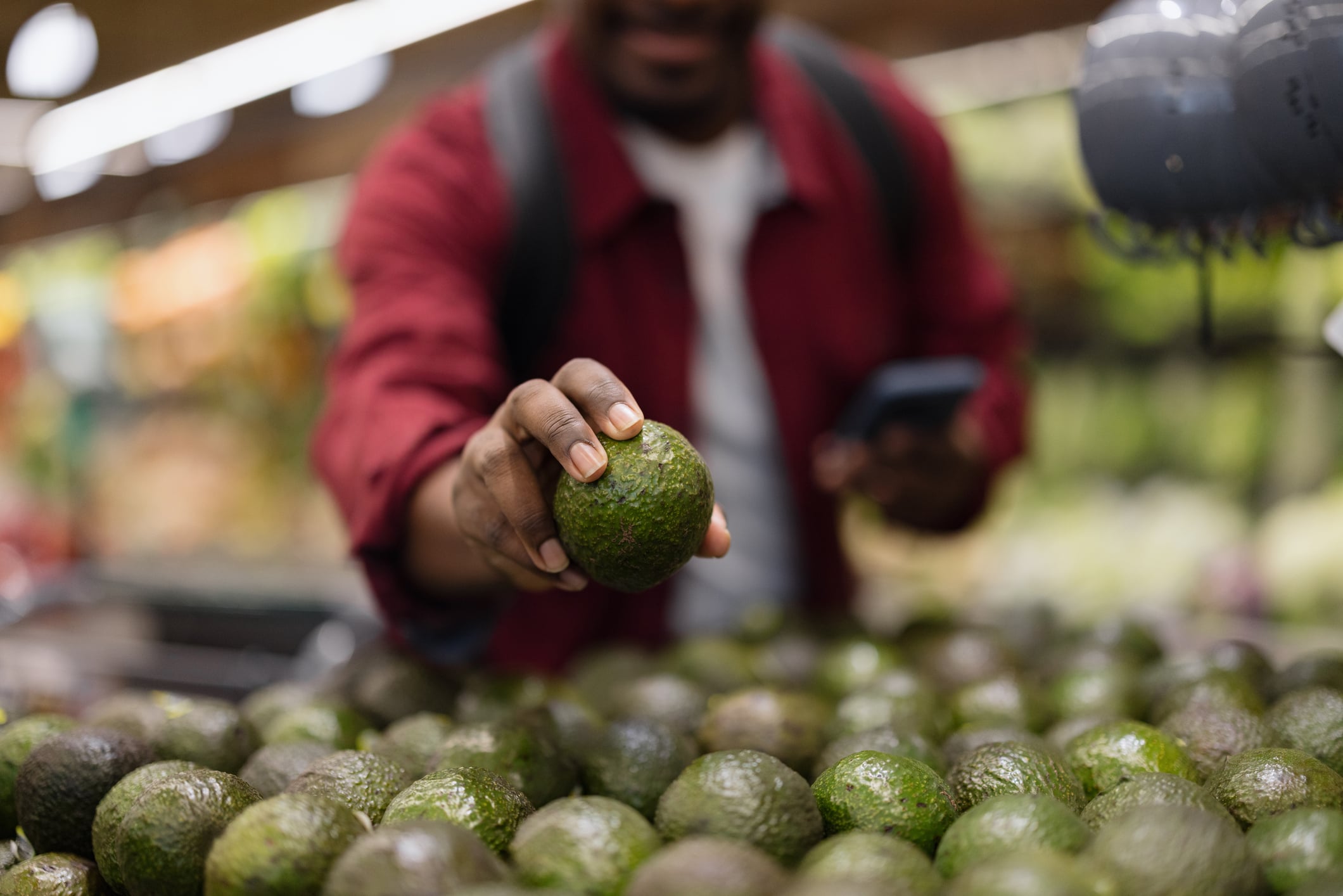 A person wearing a red jacket selects an avocado from a pile at a grocery store, holding a phone in their other hand. The background is blurred, focusing on the hand and avocados.