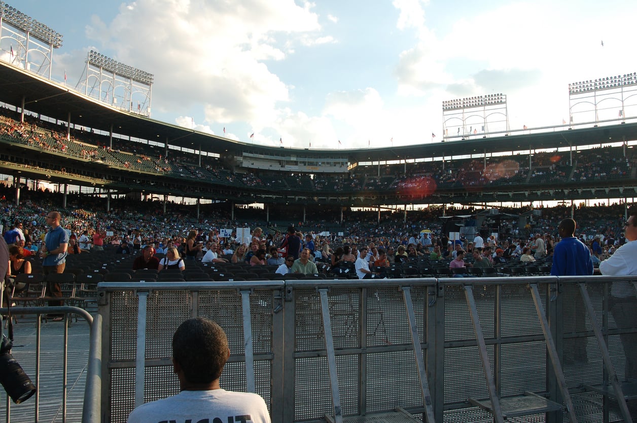A large outdoor stadium with scattered spectators in the stands and on the field under a partly cloudy sky. Metal barriers and a seated person in the foreground hint at great concert ticket hacks for accessing events like this.