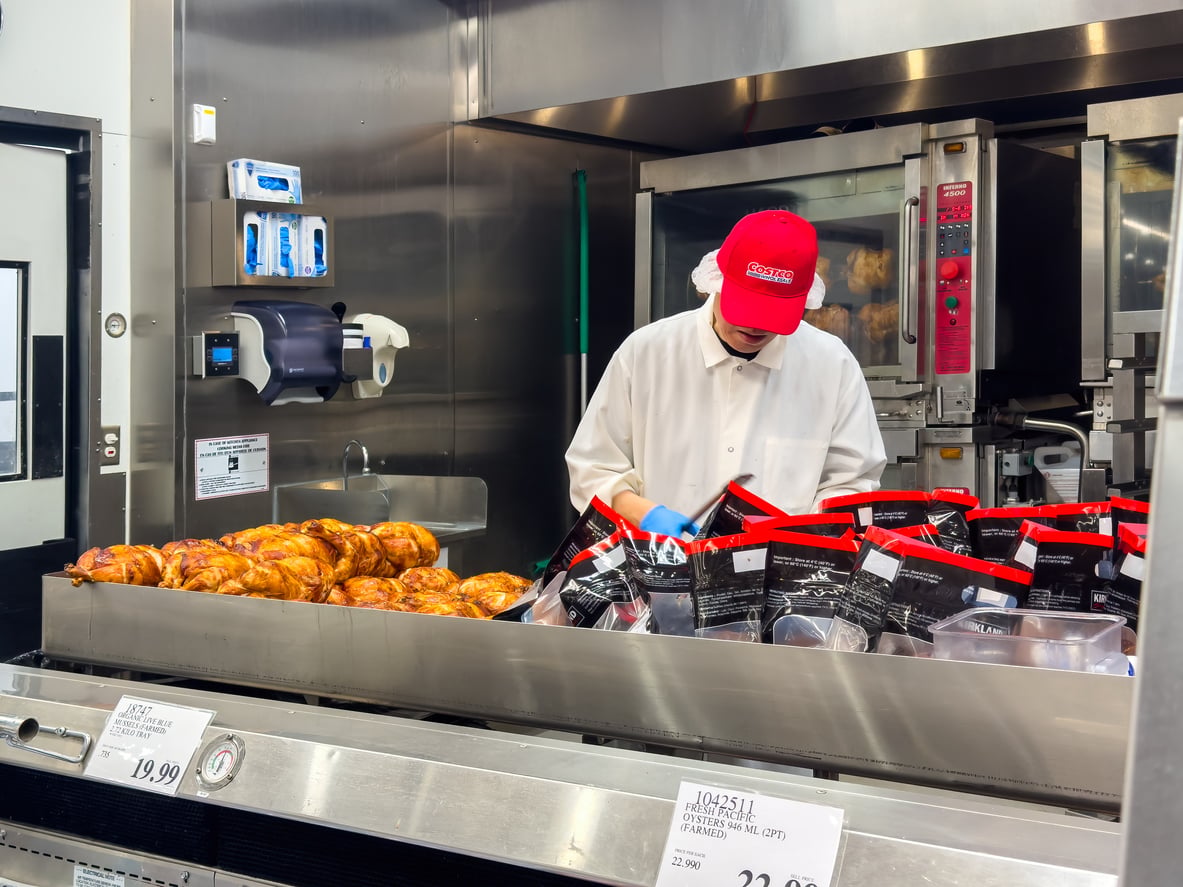 A worker in a white coat and red cap prepares and packages Costco’s famous Rotisserie Chicken, with freshly cooked chickens displayed on the left and sealed bags of rotisserie chicken being arranged on the right.