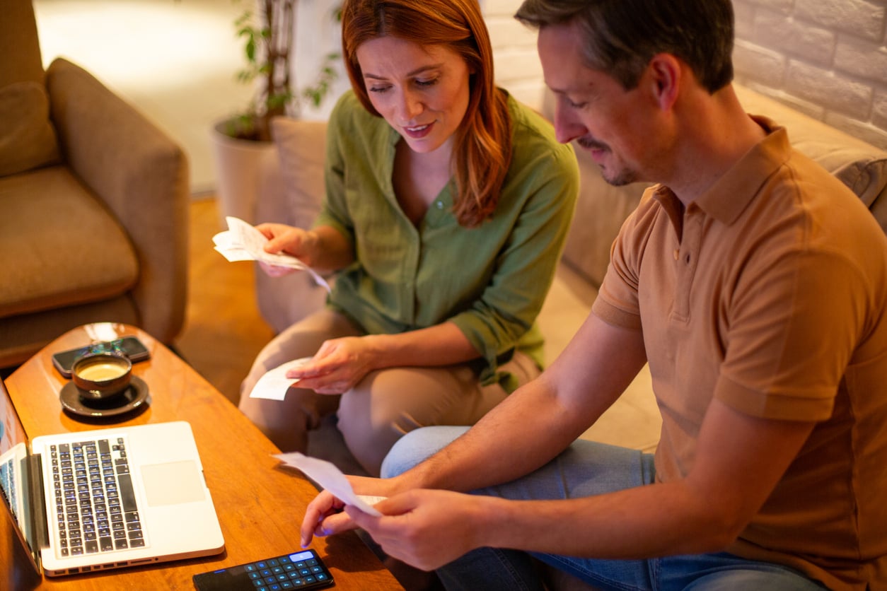 A man and woman sit together at a coffee table, reviewing receipts and using a calculator. A laptop, pen, and cup of coffee are on the table, suggesting they are managing finances at home.