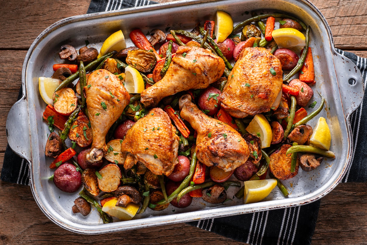 A roasting pan filled with baked chicken drumsticks, red potatoes, carrots, green beans, mushrooms, and lemon wedges—perfect for those seeking delicious ground beef alternatives—garnished with herbs on a wooden table with a striped cloth underneath.