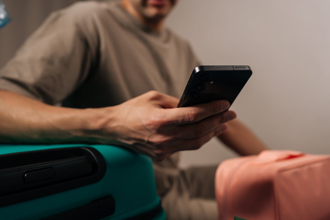 A person in a taupe shirt sits between a teal and a peach suitcase, holding and looking at a smartphone—perhaps checking Hotel Room Safety tips. The focus is on the person’s hand and phone, with their face partially out of frame.