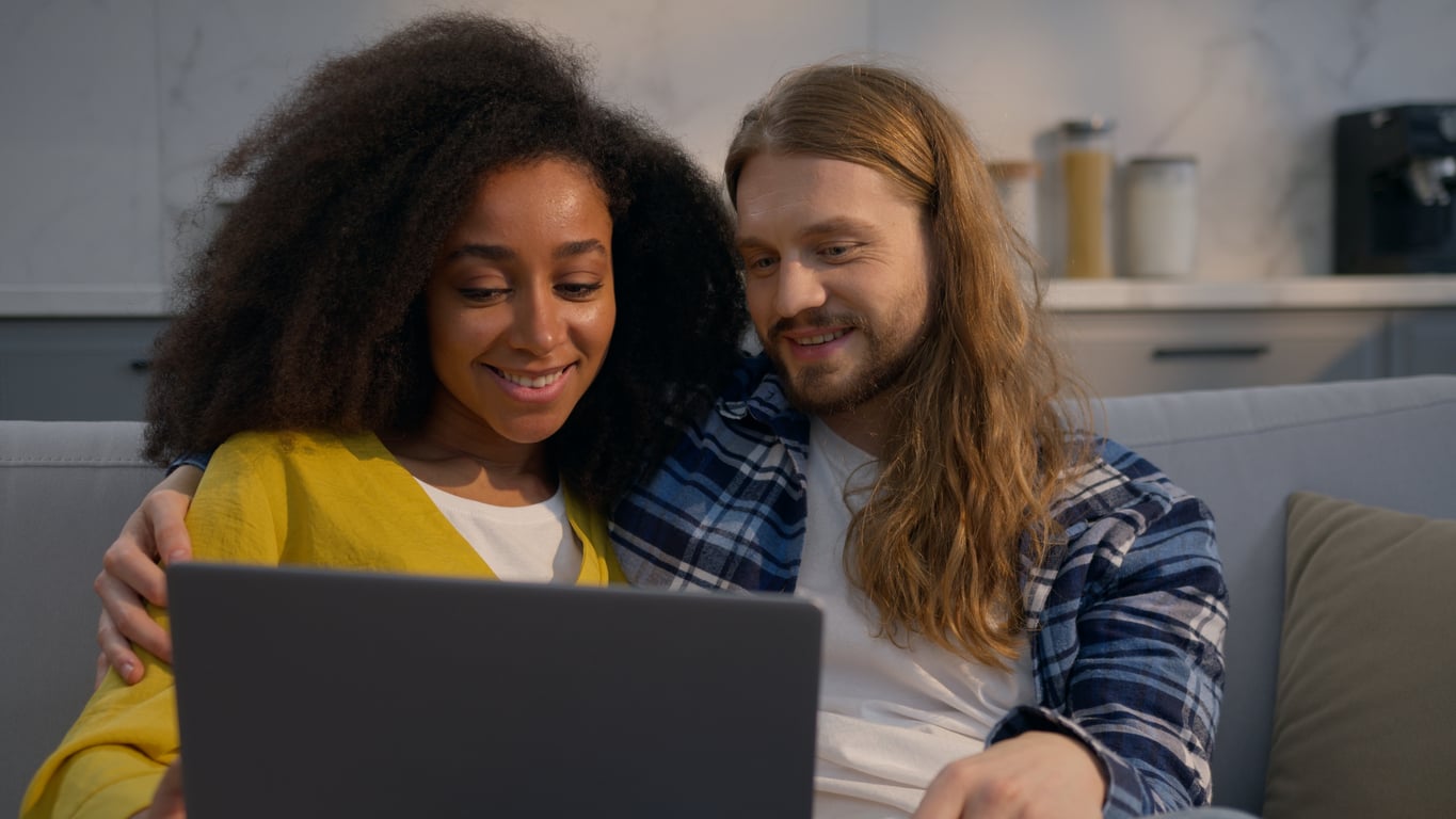 A smiling couple sits close together on a couch, looking at a laptop screen. The woman has curly hair and wears a yellow top, while the man has long hair, a beard, and wears a plaid shirt. They appear relaxed and happy.