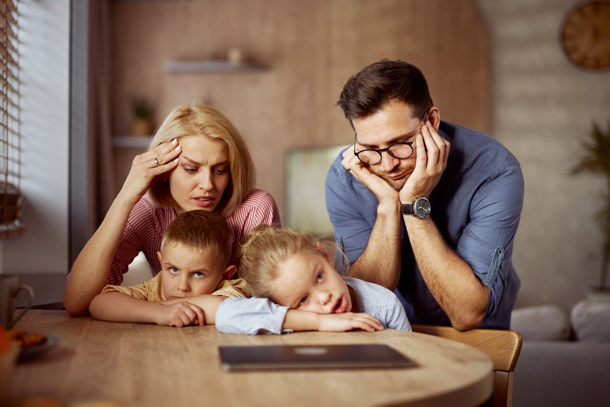 A frustrated family of four sits at a table, stressed and bored while staring at a laptop screen—delayed purchases have left the parents with their heads in their hands, as the children lean on the table, appearing disinterested.