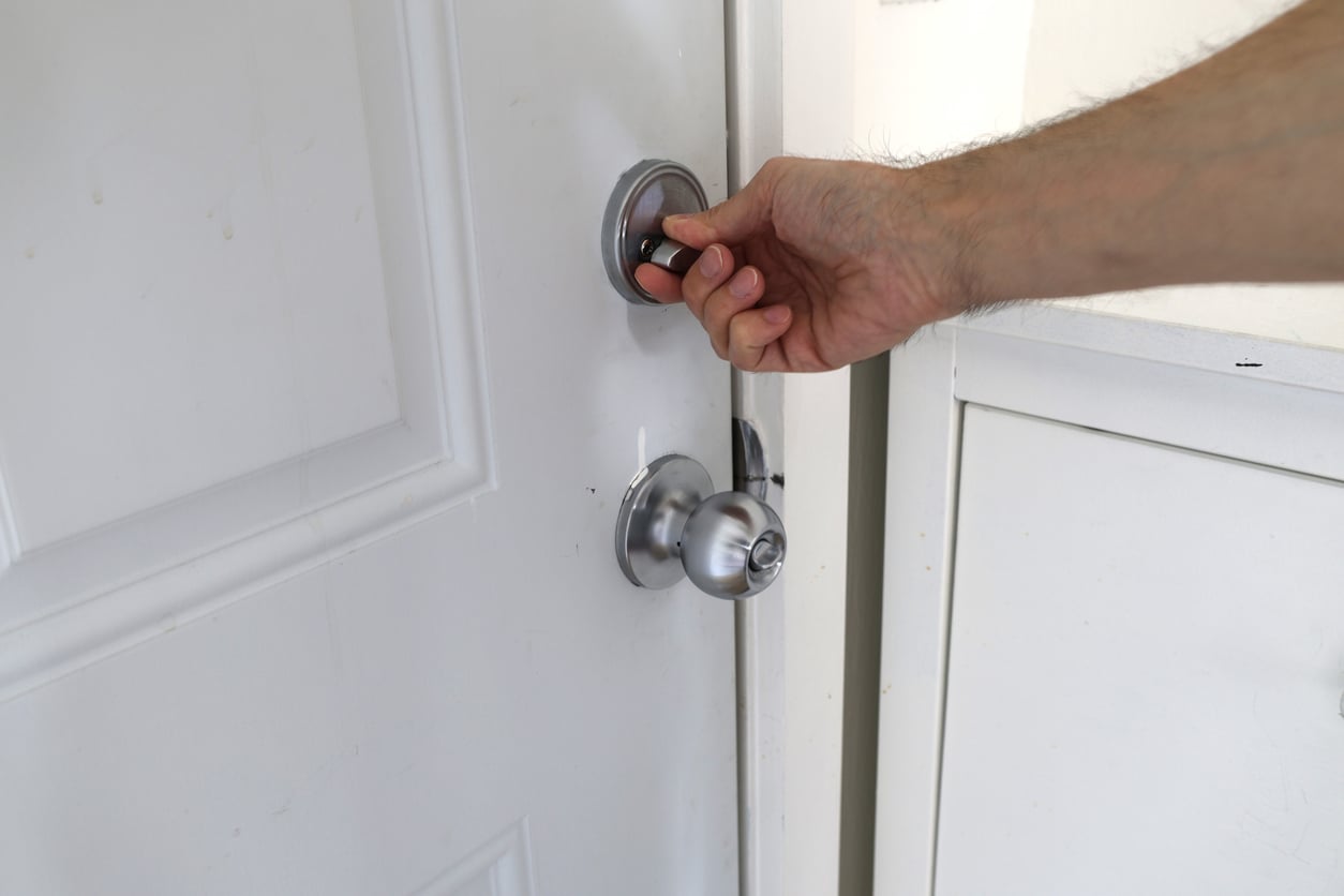 A person’s hand is shown unlocking a silver deadbolt lock on a white door above a matching door knob, highlighting Hotel Room Safety.
