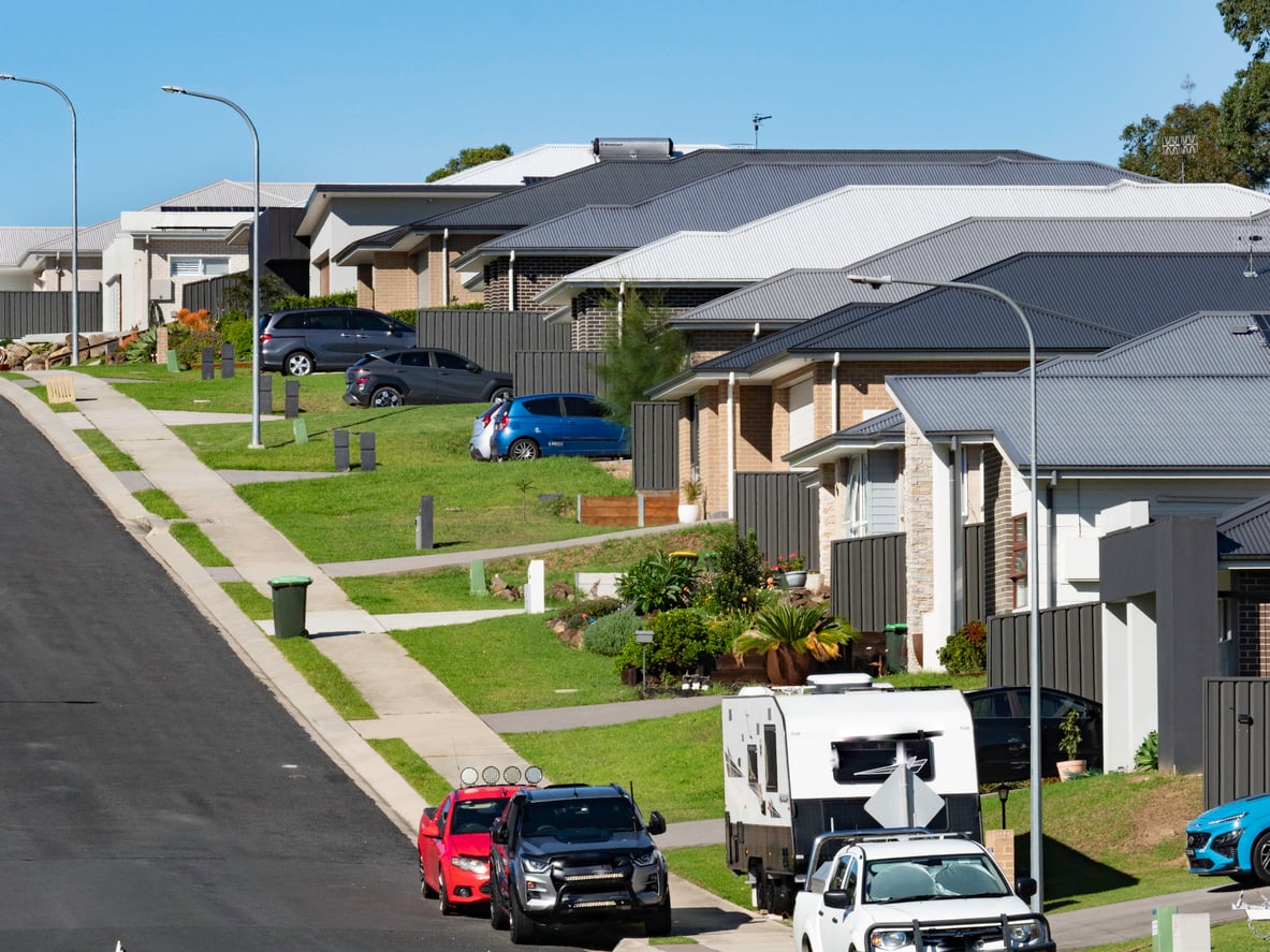 A suburban street with modern houses, neatly kept lawns, several parked cars, a trash bin, and a white RV along the curb on a clear, sunny day.