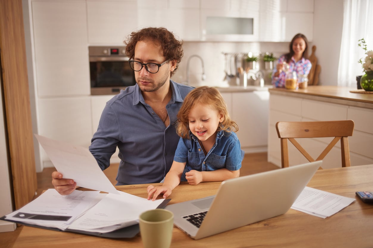 A man sits at a kitchen table with a laptop and papers, reviewing documents while a young child smiles beside him. A woman stands in the background, smiling near the counter. The kitchen is bright and modern.