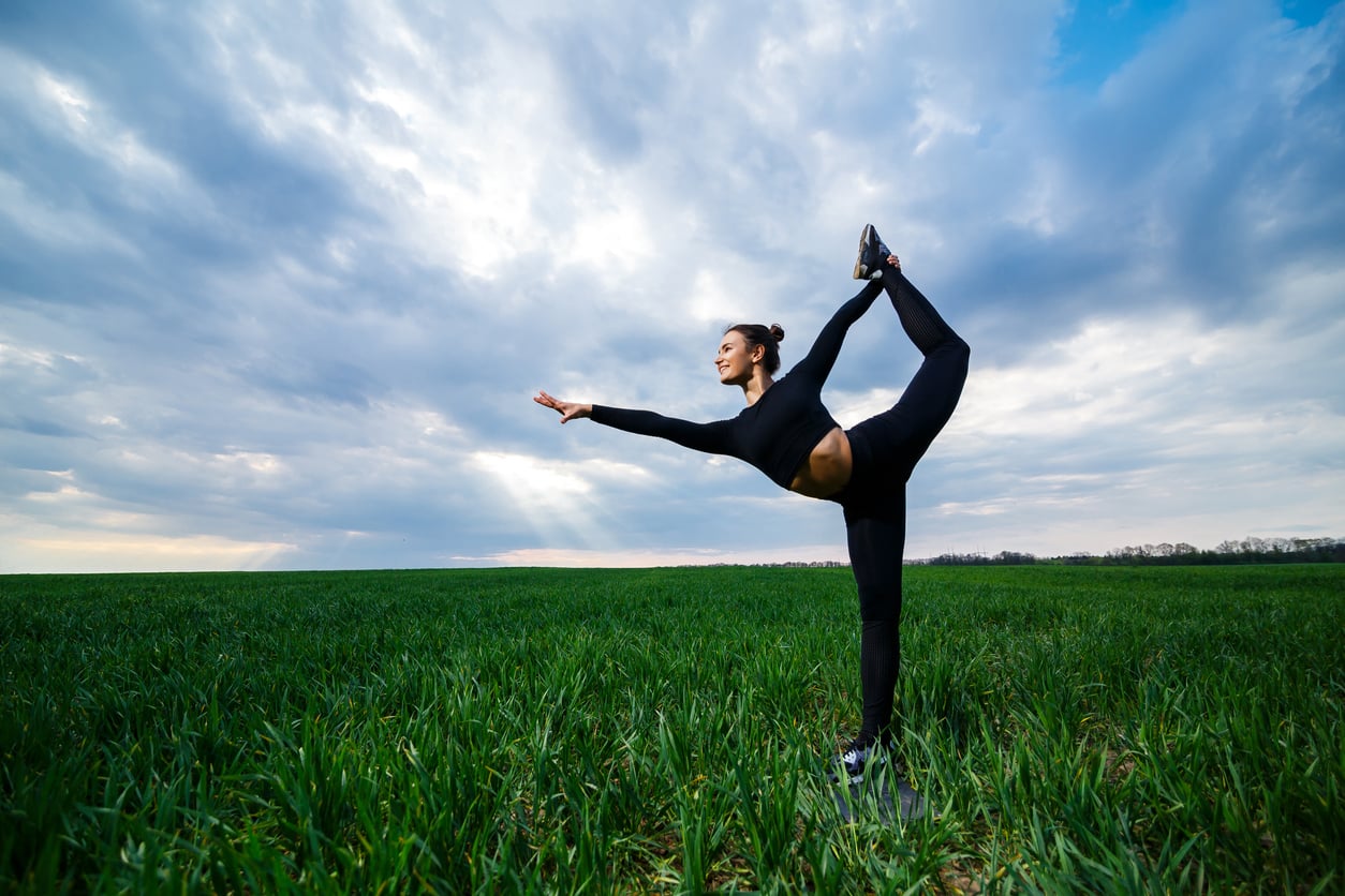 A person in black athletic wear performs a standing yoga pose in a green field under a cloudy sky with sun rays breaking through, embracing the benefits of yoga for physical fitness and mental health.