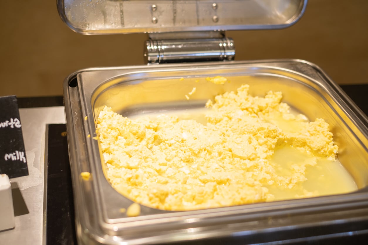 A stainless steel buffet tray with scrambled eggs in a serving line, partially submerged in liquid, with the lid open. A small blackboard sign is visible on the side.