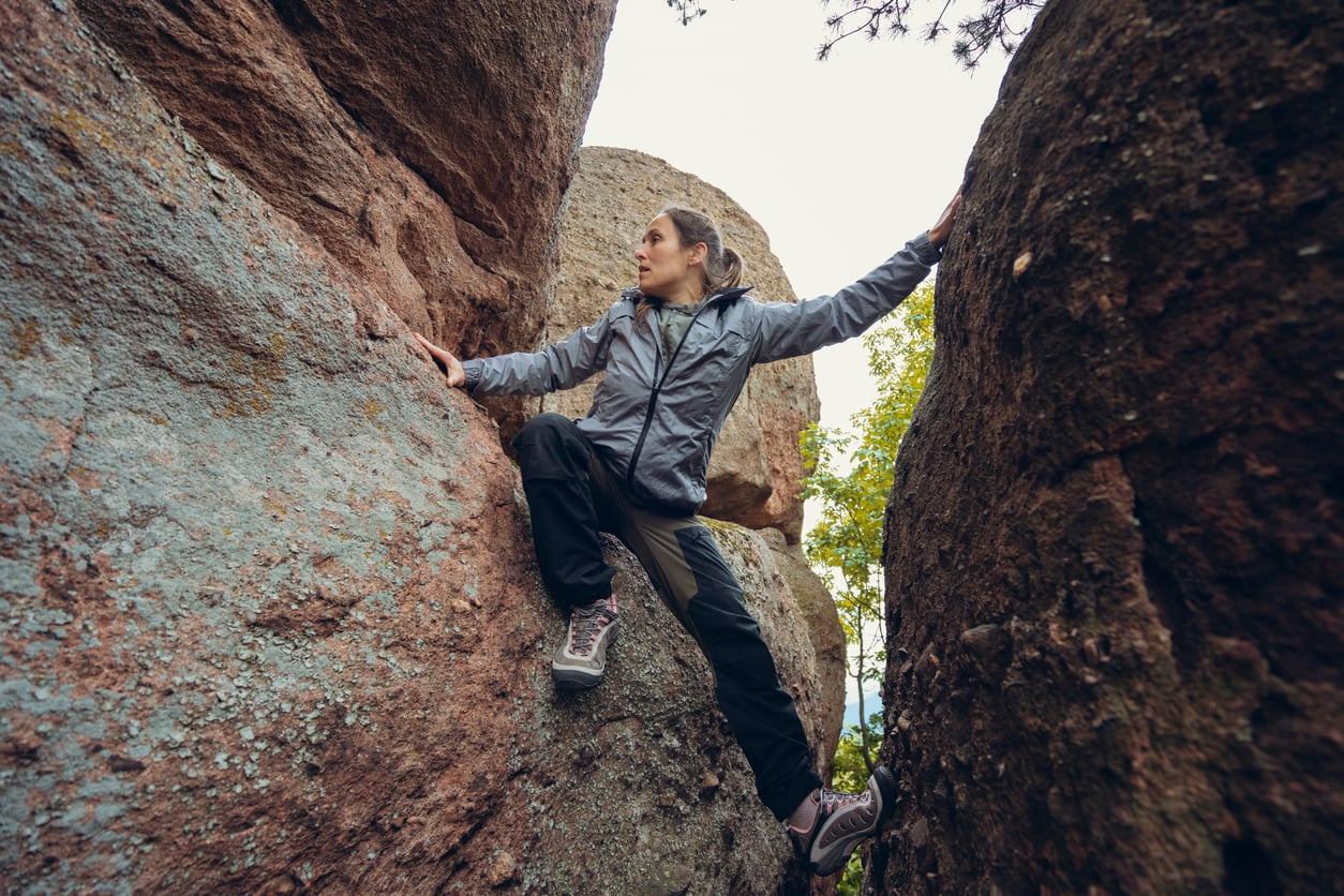 A person wearing a gray jacket and hiking boots climbs between two large rock formations, using hands and feet to balance—showing that exciting Florida alternatives for outdoor adventure await beyond the usual beaches. Trees and sky fill the background.