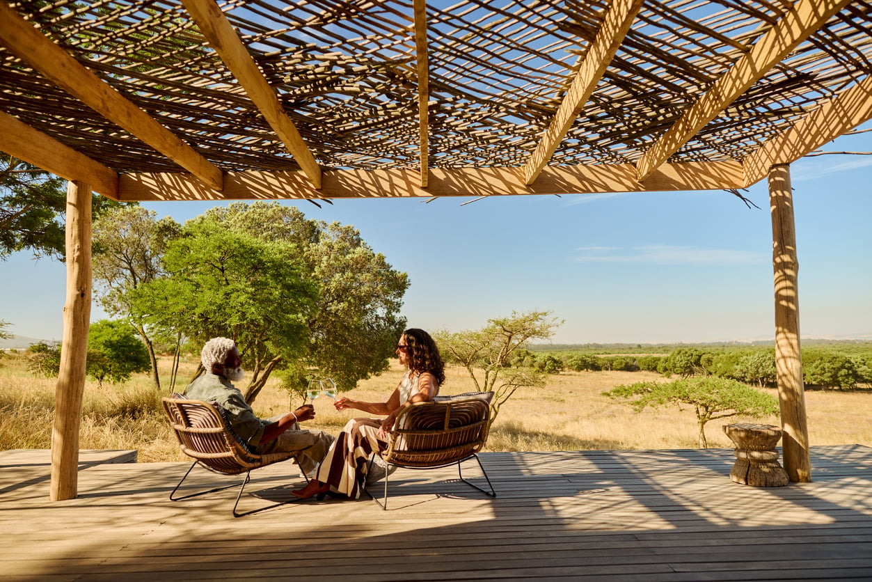 Two people sit on wicker chairs under a wooden pergola, enjoying drinks and facing a scenic, sunlit savanna landscape with trees and open grasslands.