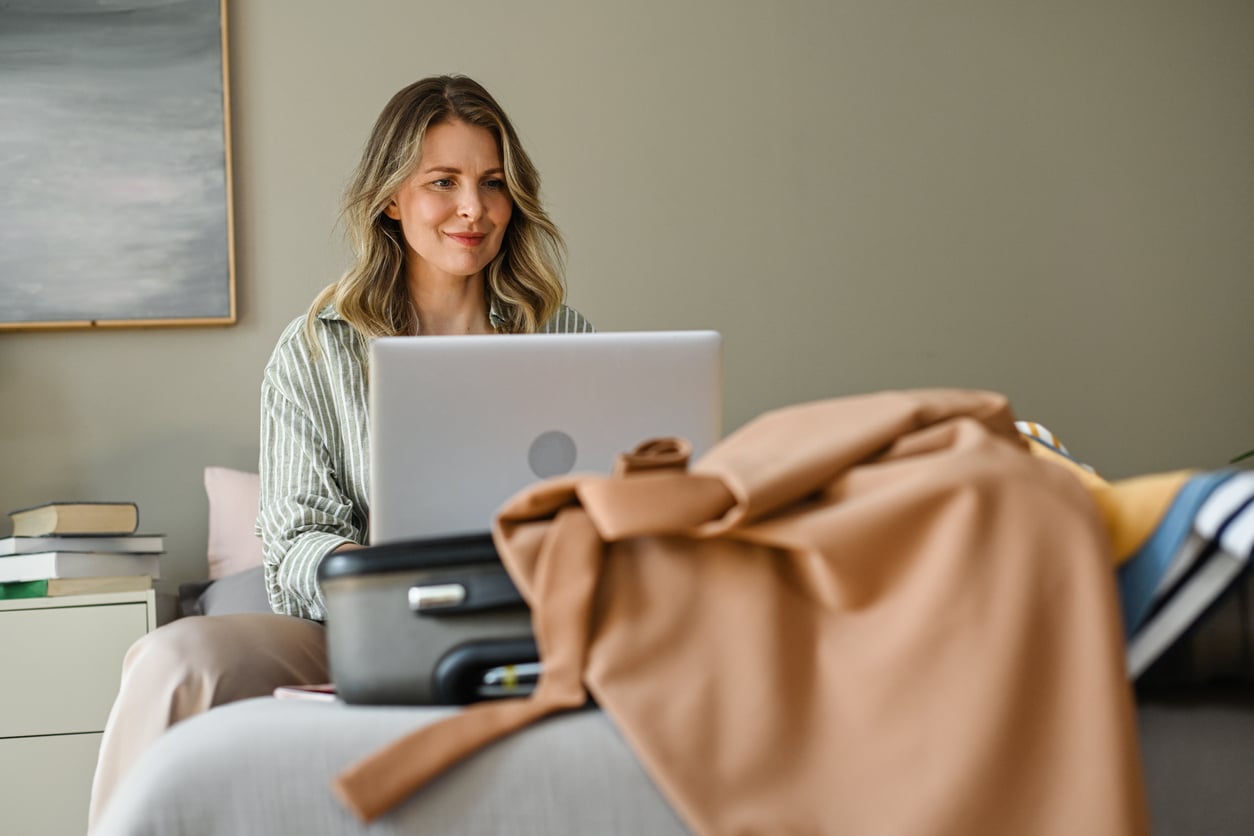 A woman sits on a bed using a laptop, with an open suitcase and clothes beside her. She appears to be packing or working while preparing for a trip. There are books and a painting in the background.