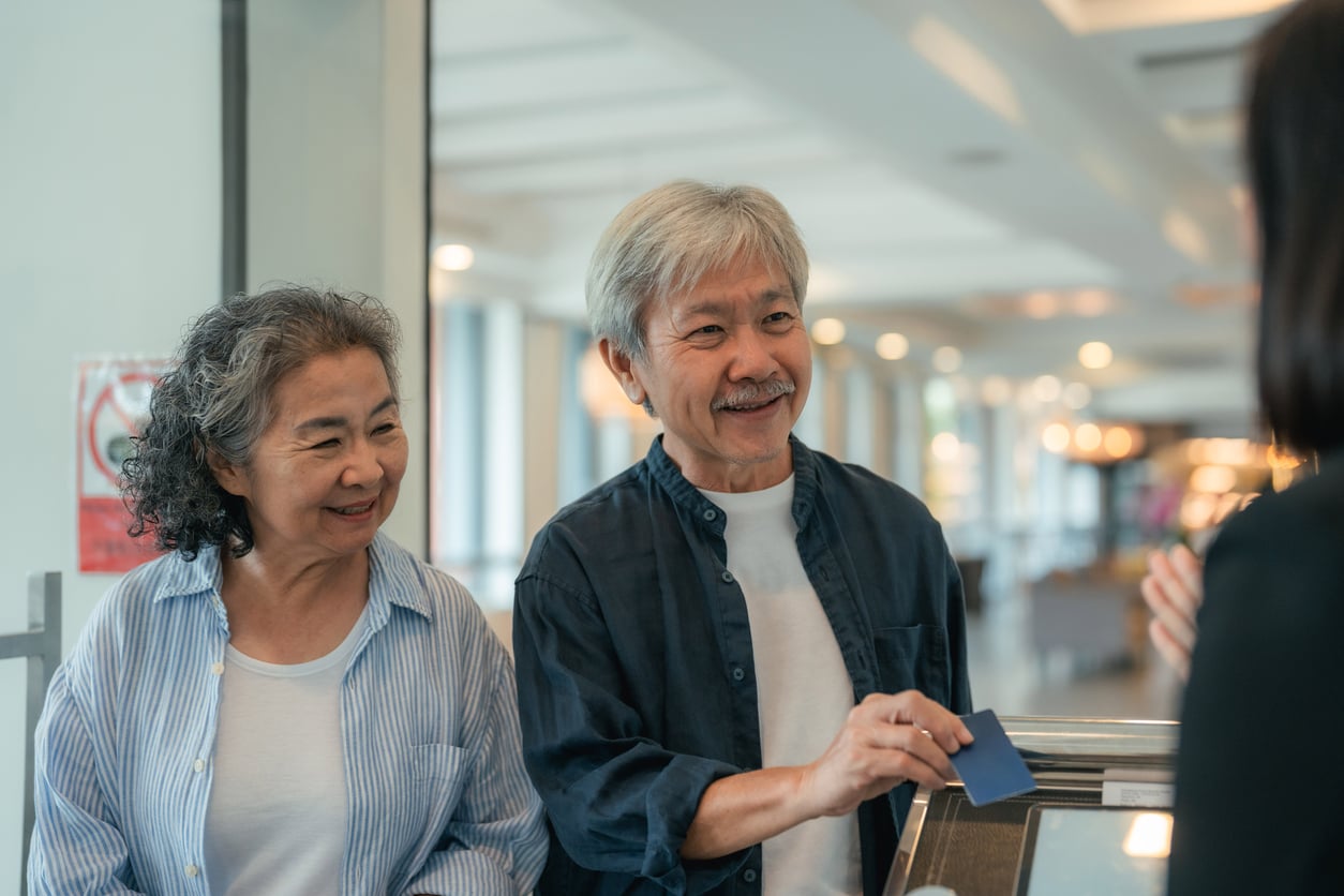 An older couple stands at a counter smiling, with the man holding a blue card—possibly using travel hacks while making a payment or checking in—chatting with someone across from them in a brightly lit indoor setting.