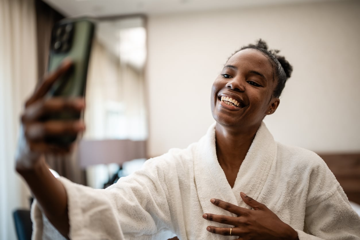 A smiling woman in a white bathrobe takes a selfie with her smartphone in a bright, modern hotel room, holding one hand to her chest and looking happy, enjoying the comfort and hotel room safety during her stay.