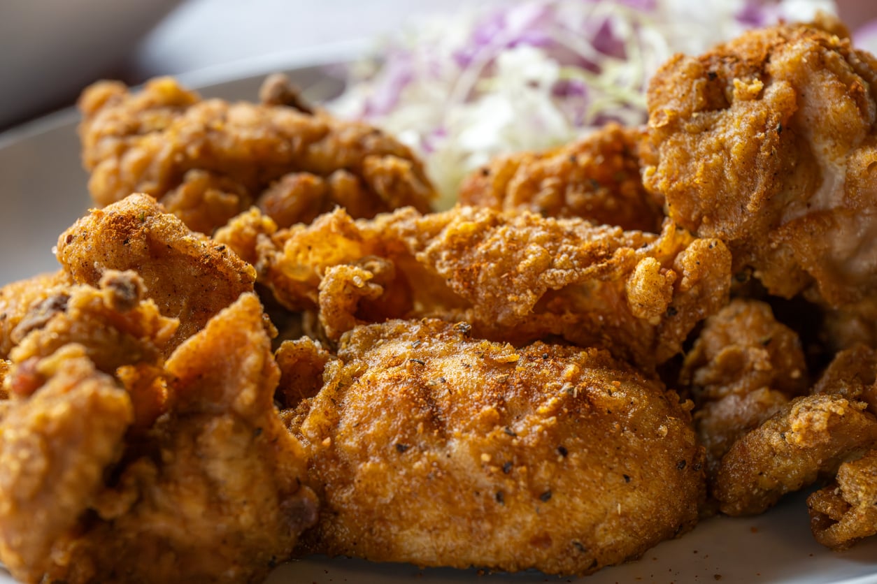 Close-up of crispy, golden-brown fried chicken pieces on a plate, with a side of shredded cabbage and purple vegetables blurred in the background.